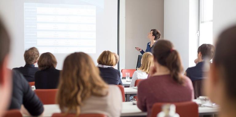 a woman gives a power point presentation to a group of people sitting in desks.