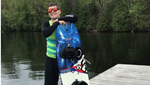 Josh is standing on the edge of the dock wearing a black shirt and shorts with a green life jacket. A set of blue and yellow water skiis on the dock, and he’s leaning against a wakeboard. He’s wearing sunglasses and smiling.