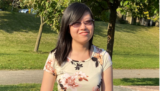 Lily is outside, standing in front of two lush green trees. She is smiling and wears a floral top and sunglasses. 