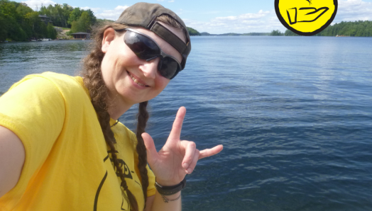 Jessica Bailey, poses at the Lake Joe waterfront. She does the American Sign Language sign for "I Love you". An icon of a hand with a heart floating above it appears in the top right hand corner. 