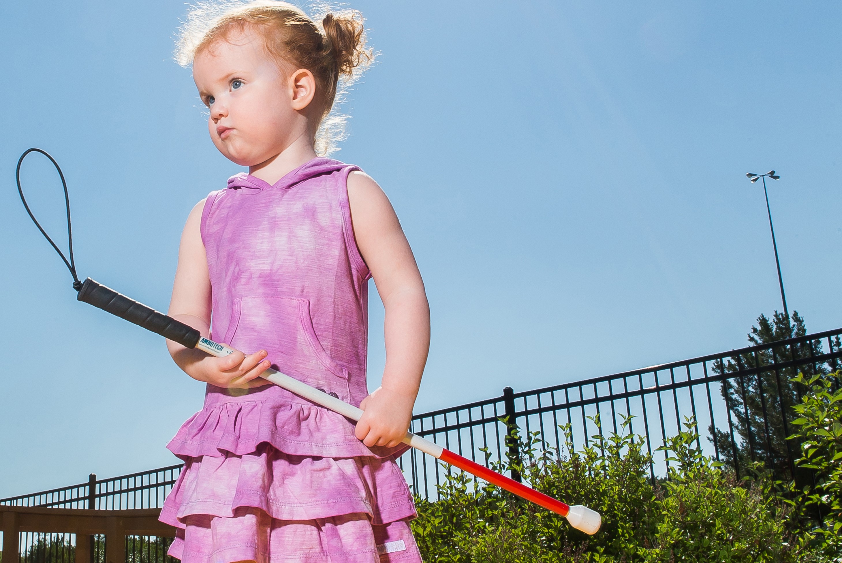 A young girl with sight loss, wearing a pink dress, holding a white cane outside,