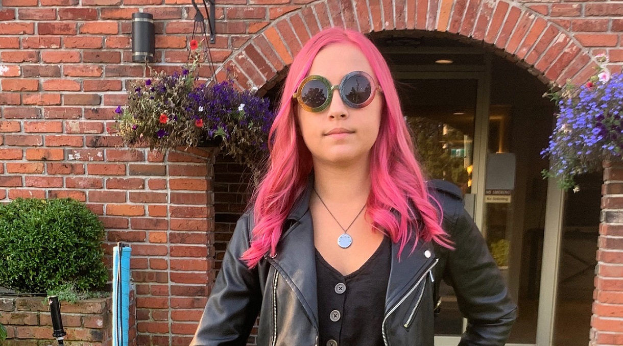 A cool teenager stands outside her house with her white cane in hand. She has pink hair and is wearing a leather jacket.