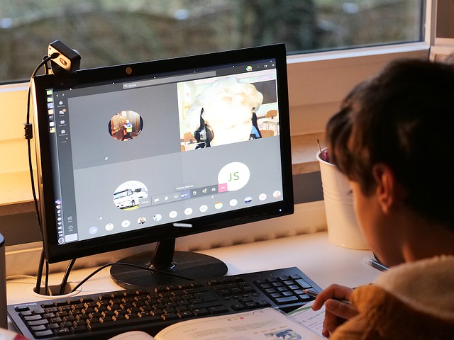 A young child sits at a desktop computer. The computer screen displays a Zoom call.