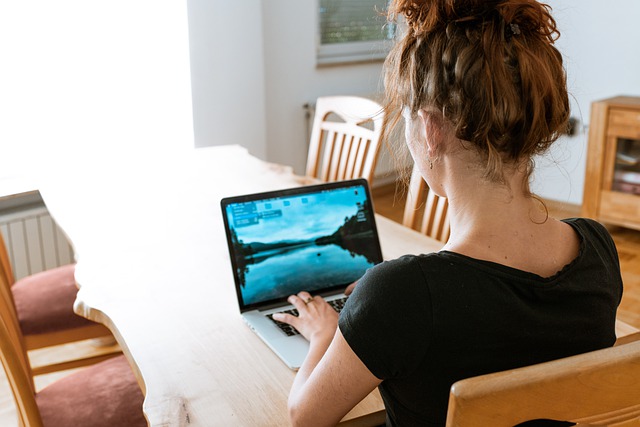 The back of a young woman's head. She sits at kitchen table and types on her laptop computer.