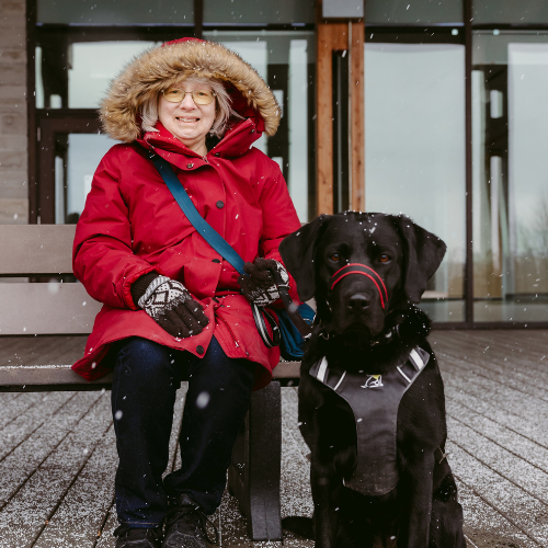 À l’extérieur, Cheri portant un blouson d’hiver rouge et des gants sourit, assise sur un banc, et son chien-guide, Sassy, une chienne noire munie d’un harnais est assis sur le sol à sa droite.