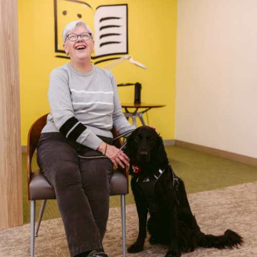 Dans un bureau d’INCA, Penny est assise sur une chaise. Son chien-guide, Honour, un croisement de Labrador Retriever noir et de Golden Retriever, est assis à ses pieds. Penny caresse la tête d’Honour et sourit de joie. 