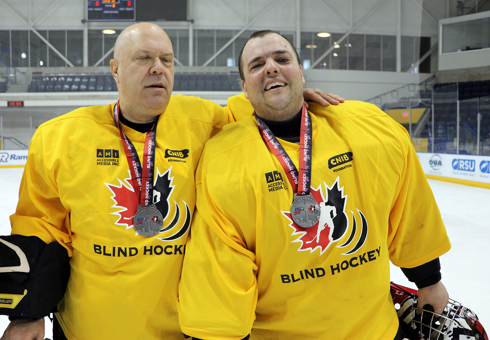 Deux hommes adultes, gardiens de but de hockey sonore, se tiennent sur la glace côte à côte, portant des chandails, des jambières et une médaille autour du cou. 