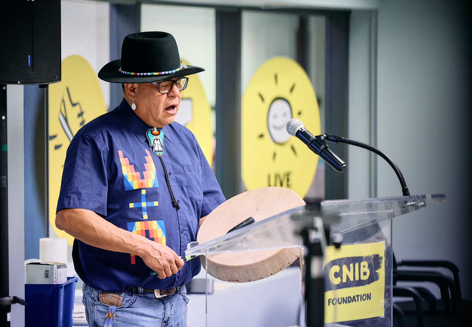 Un Autochtone chante en jouant du tambour, debout sur un podium portant le logo d'INCA. 