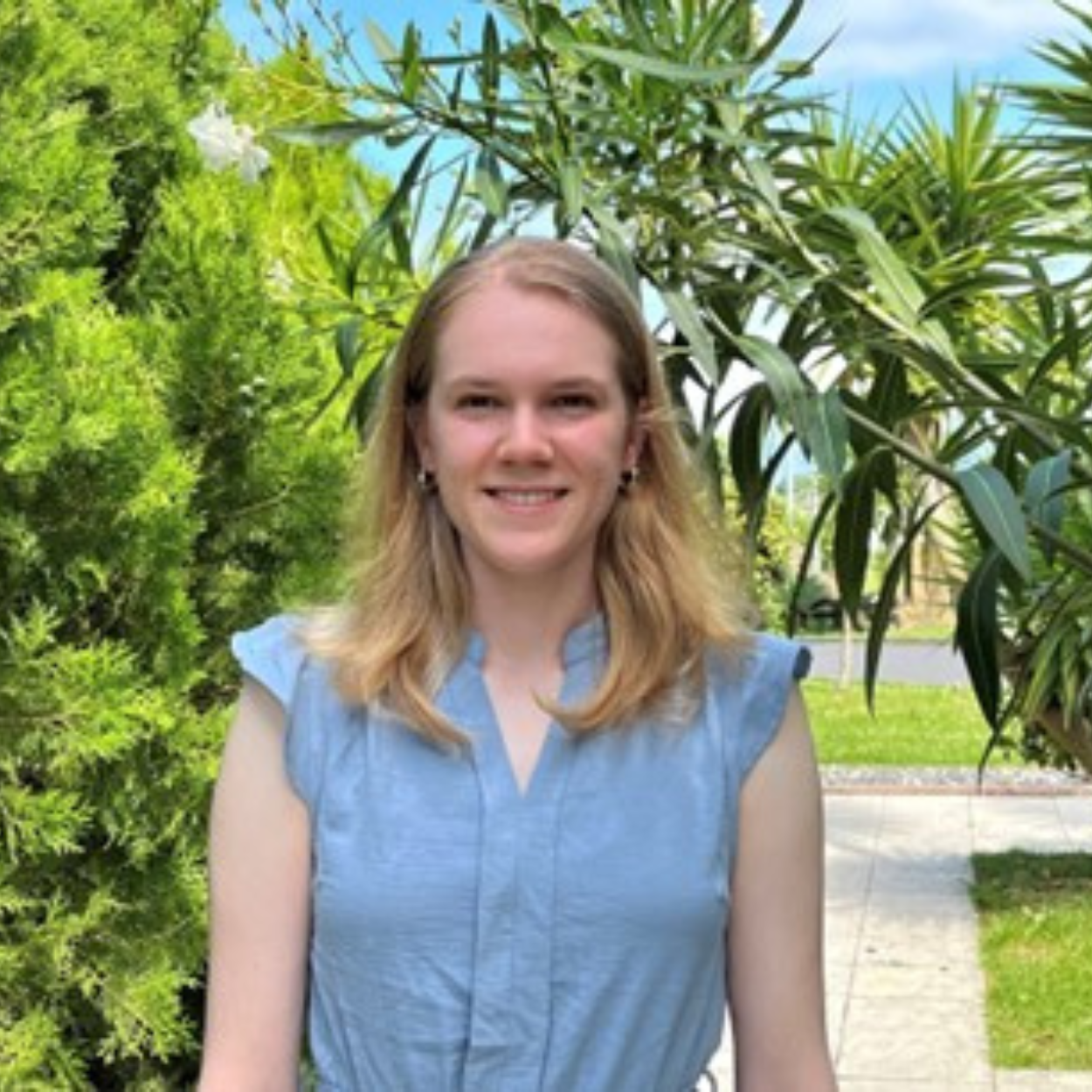 Laura, une jeune femme aux cheveux blonds, sourit devant un jardin verdoyant avec des palmiers et des arbustes. Elle porte une robe de soleil bleu clair.