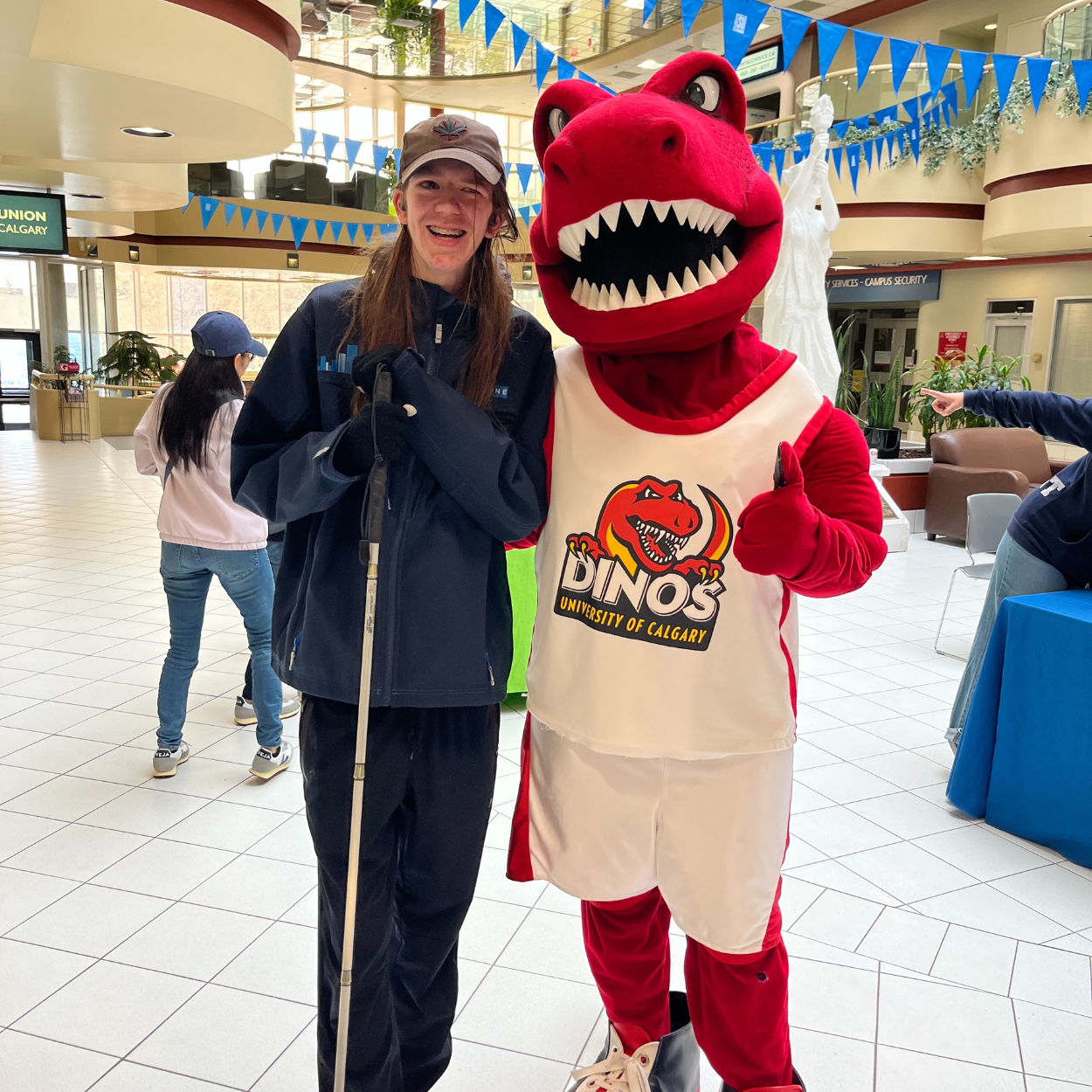 Zachary, un jeune homme aux longs cheveux bruns, sourit à côté de Rex, la mascotte dinosaure rouge et duveteuse de l'université de Calgary. Zach est vêtu d'un sweat-shirt bleu, d'un pantalon de survêtement, d'un chapeau et tient sa canne blanche.