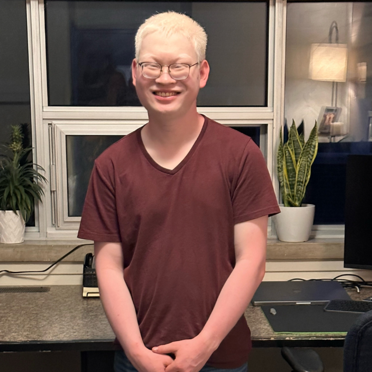 Aidan, un jeune homme aux cheveux blancs, se tient devant un bureau à domicile/un poste de travail informatique. Aidan porte des lunettes et un t-shirt marron avec un jean. Il sourit et se tient debout, les mains jointes.