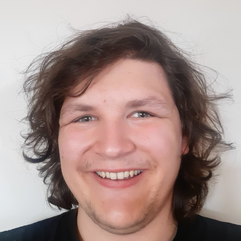 Tate, un jeune homme aux cheveux bruns, pose pour une photo de type selfie. Il porte un t-shirt noir et sourit.