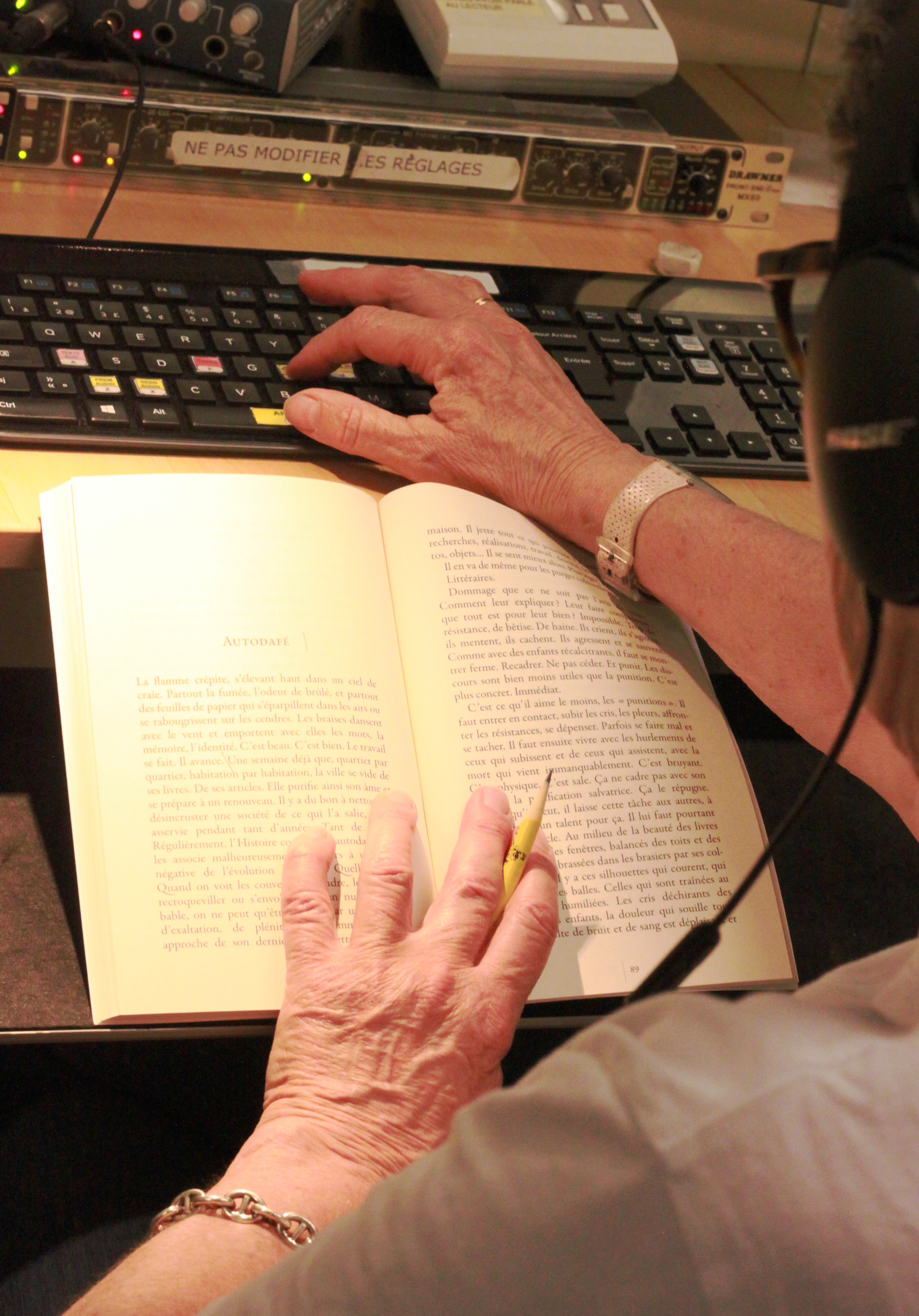 Gros plan d'une femme devant un livre, crayon à la main, écouteur sur les oreille et sa seconde main sur un clavier devant des outils d'enregistrement