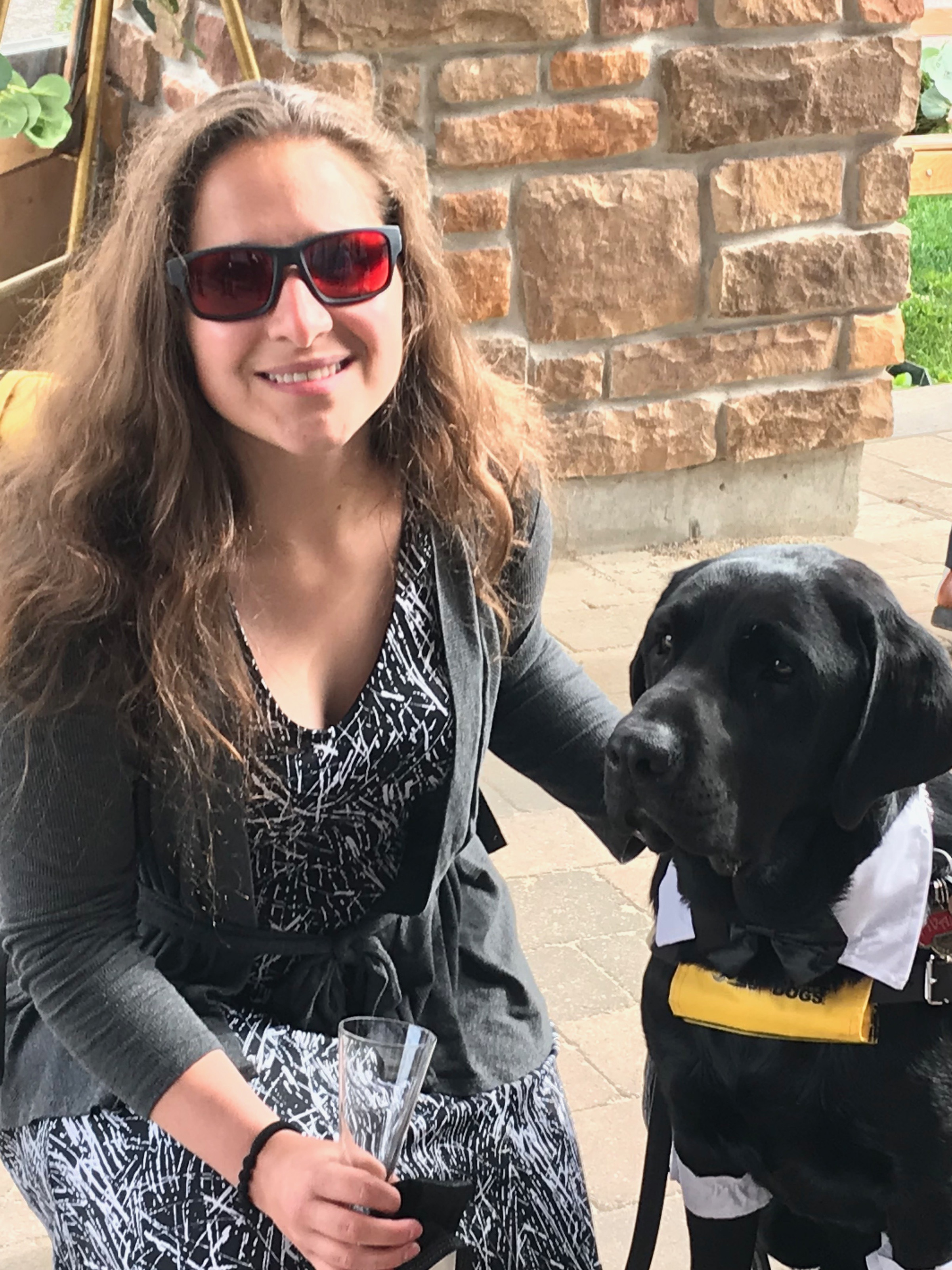 Amanda, kneeling outside wearing red tinted sunglasses, posing for the camera next to Ivan, her CNIB Guide Dog. 