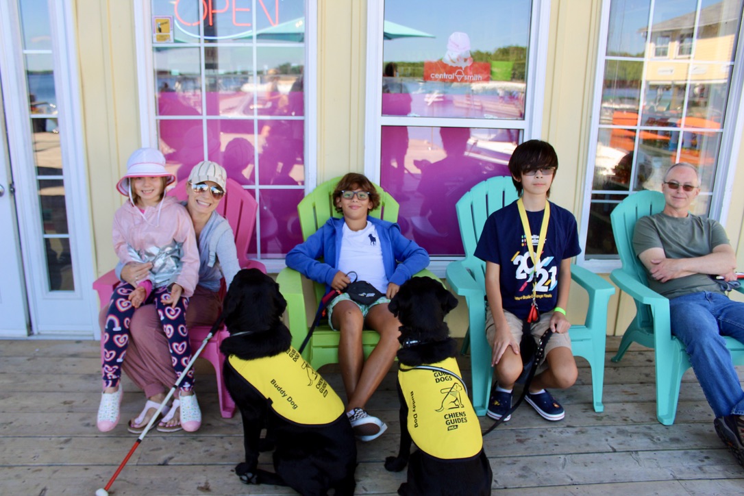 12-year-old Gabriel sits in a colouful Muskoka chair, relaxed and smiling, surrounded by other happy CNIB Lake Joe campers and families, and two CNIB buddy dogs in yellow vests.