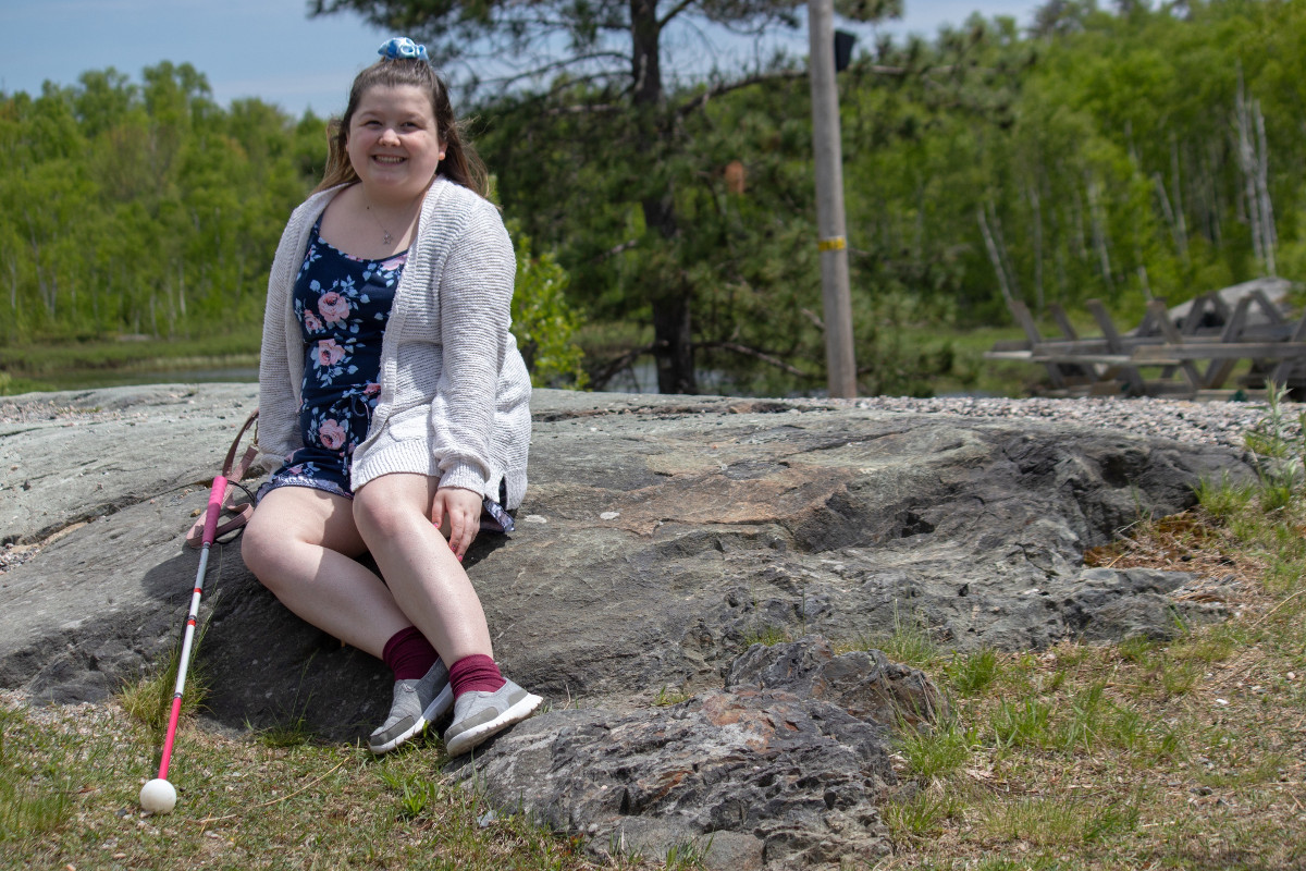 Alicia smiles and sits outdoors on a large rock. Behind her are trees. She is wearing a summer floral dress. Her white cane rests on the rock to the right of her.