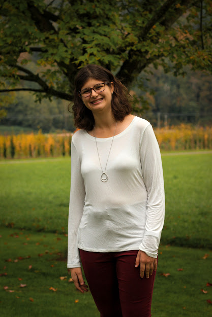 Amy Verwoerd pose à l’extérieur devaAmy Verwoerd poses for a photo outdoors with a lush green landscape of trees and grass in the background.