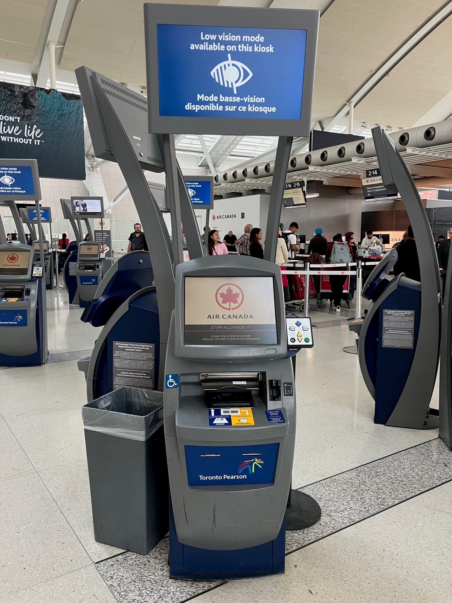 Un kiosque d'enregistrement libre-service à l'aéroport Pearson. L'écran du kiosque affiche le logo d'Air Canada et, au-dessus, un panneau indique : «  Mode basse vision disponible sur ce kiosque ». Un panneau avec des boutons de navigation accessibles est situé sur le côté droit de la borne, à côté de l'écran tactile. 