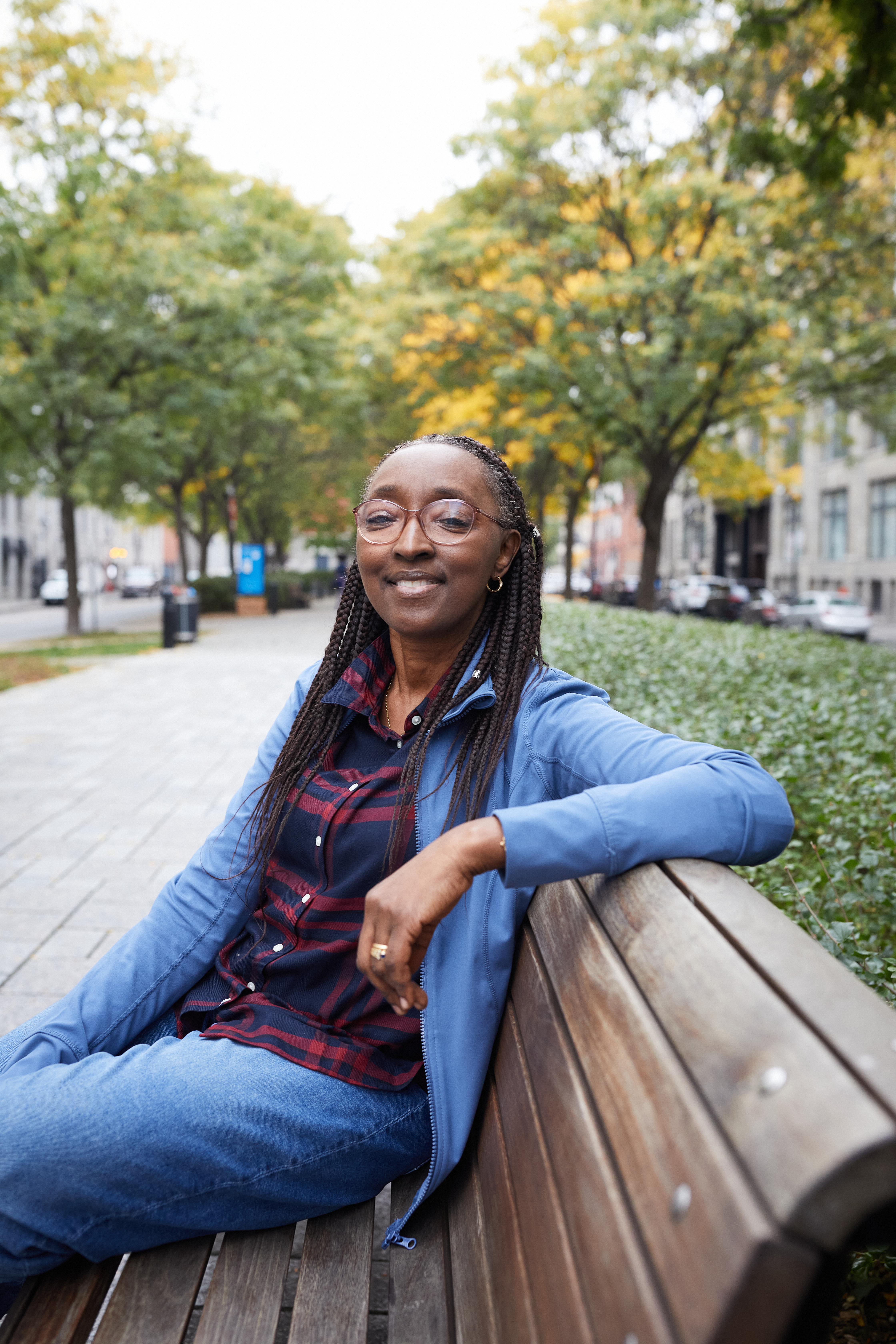 Binta is sitting on an outdoor wooden bench. She is smiling and wearing glasses.