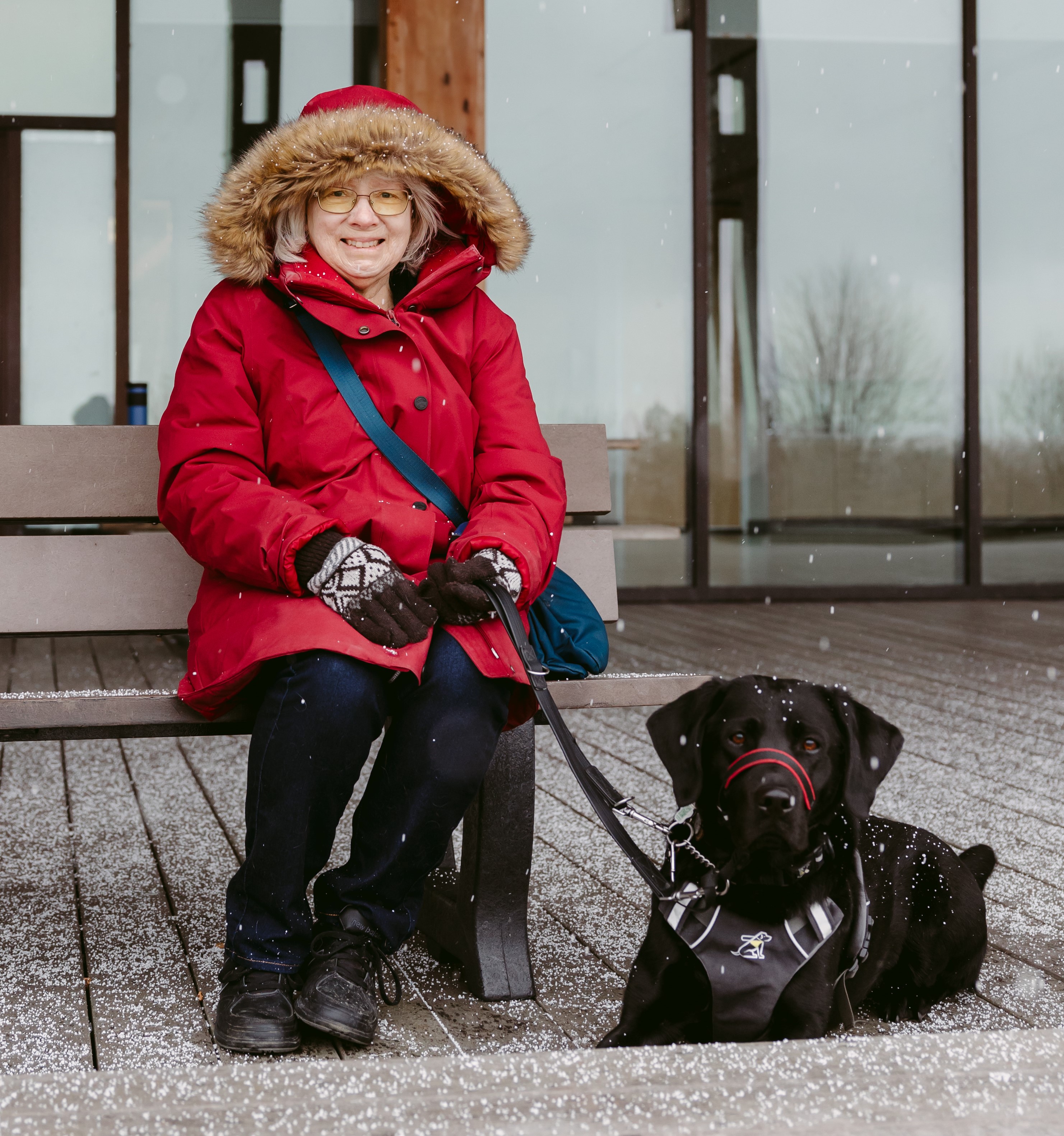 Cheri, qui porte une parka rouge à capuche, est assise sur un banc en bois avec Sassy, sa chienne-guide Labrador noire d'INCA, à ses côtés par une journée enneigée.