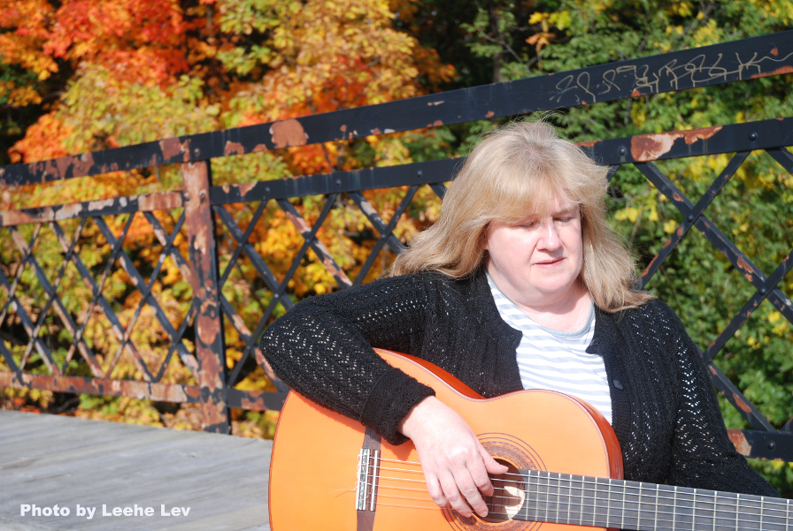 Photo of Christine playing guitar on a bridge. Trees are behind her and the sun shining. 