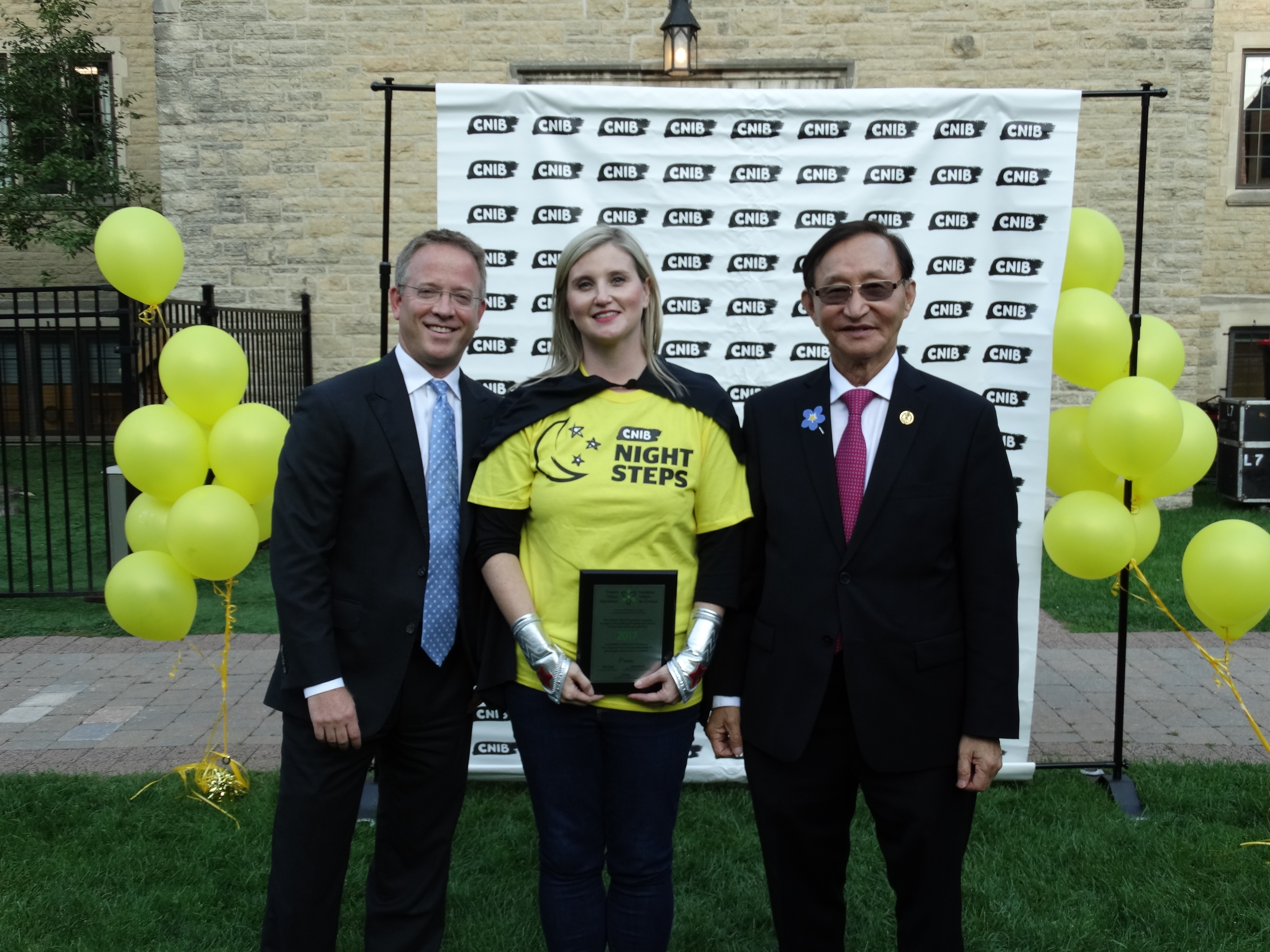 Justin Mooney, Suzanne Decary-Van den Broek and Minister Raymond Cho pose in front of a CNIB branded backdrop. Suzanne is holding a plaque. 