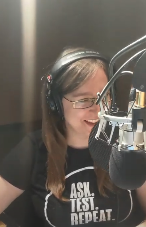 Dana sits in a recording booth. She is wearing big headphones and speaking into a microphone. 