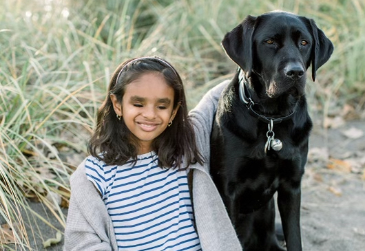 Deepa and Chelsey, a black Labrador-Retriever, sitting on sand in front of a grassy area; Deepa’s arm is around Chelsey and she is smiling for the camera.