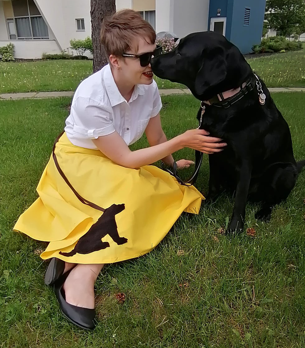Denise, sitting in the grass wearing a bright yellow skirt, bumping noses with Tara, her CNIB Guide Dog.
