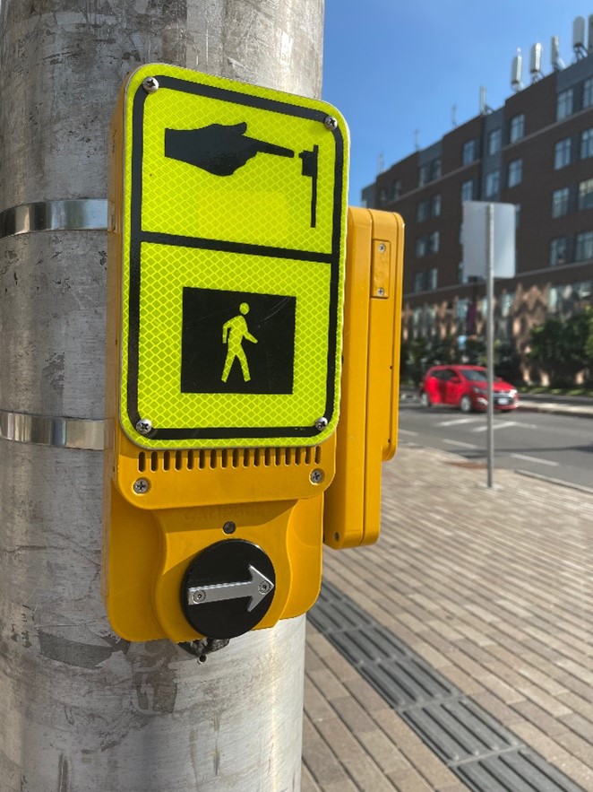 Une signalisation piétonnière accessible  monté sur un poteau métallique à un passage pour piétons. Le dispositif est de couleur jaune et se compose de plusieurs éléments.