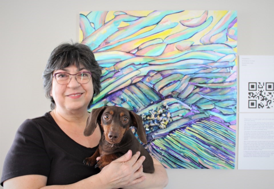 Robyn Rennie poses for a photo in front of her colourful artwork next to a digital printed QR Code. She is holding a small, chocolate-brown dog. 