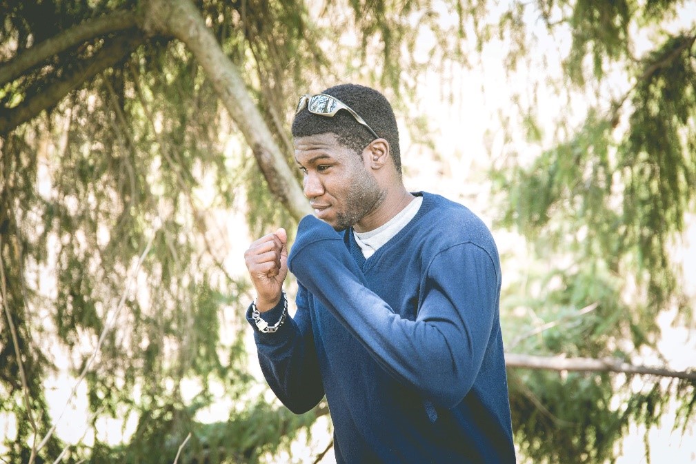 Dressed casually in jeans and a blue shirt, Bernard, strikes a boxing pose. His fists are up near his chest ready to punch. Behind him is a landscape of sweeping green trees. 