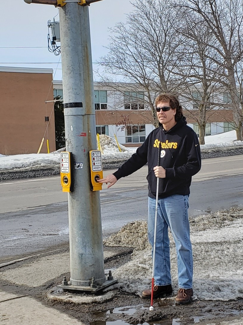 Bill Liggins stands on the corner at an Accessible Pedestrian Signal. He is holding his white cane in his left hand and pressing the crosswalk button with this right hand. 