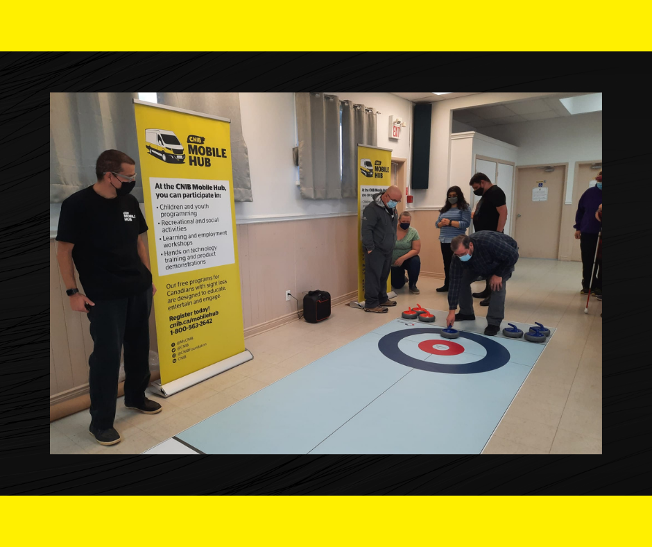 Six people play a game of floor curling inside a rec centre at the CNIB Mobile Hub Hamilton stop. 