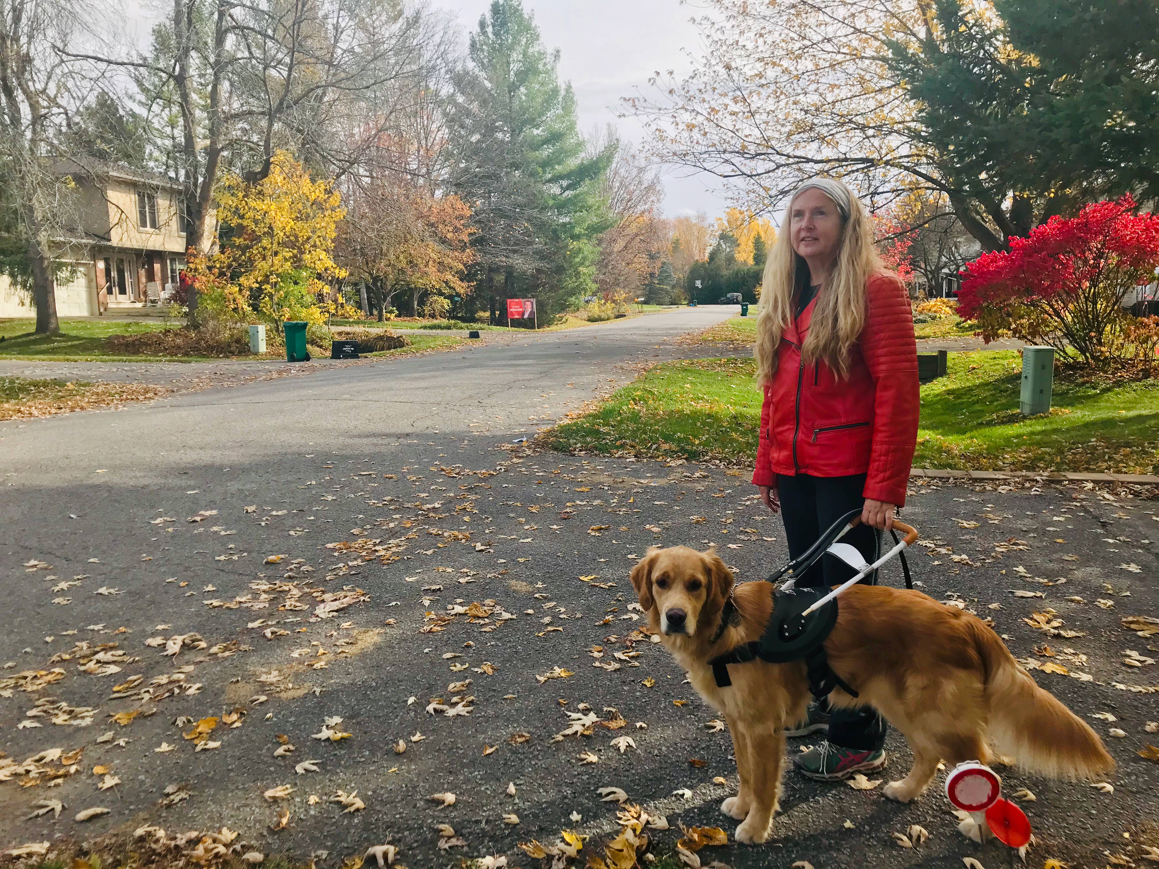 Diane, wearing a red jacket standing in a street that’s covered with colourful, autumn leaves, holding onto Carla’s harness while smiling for the camera. 