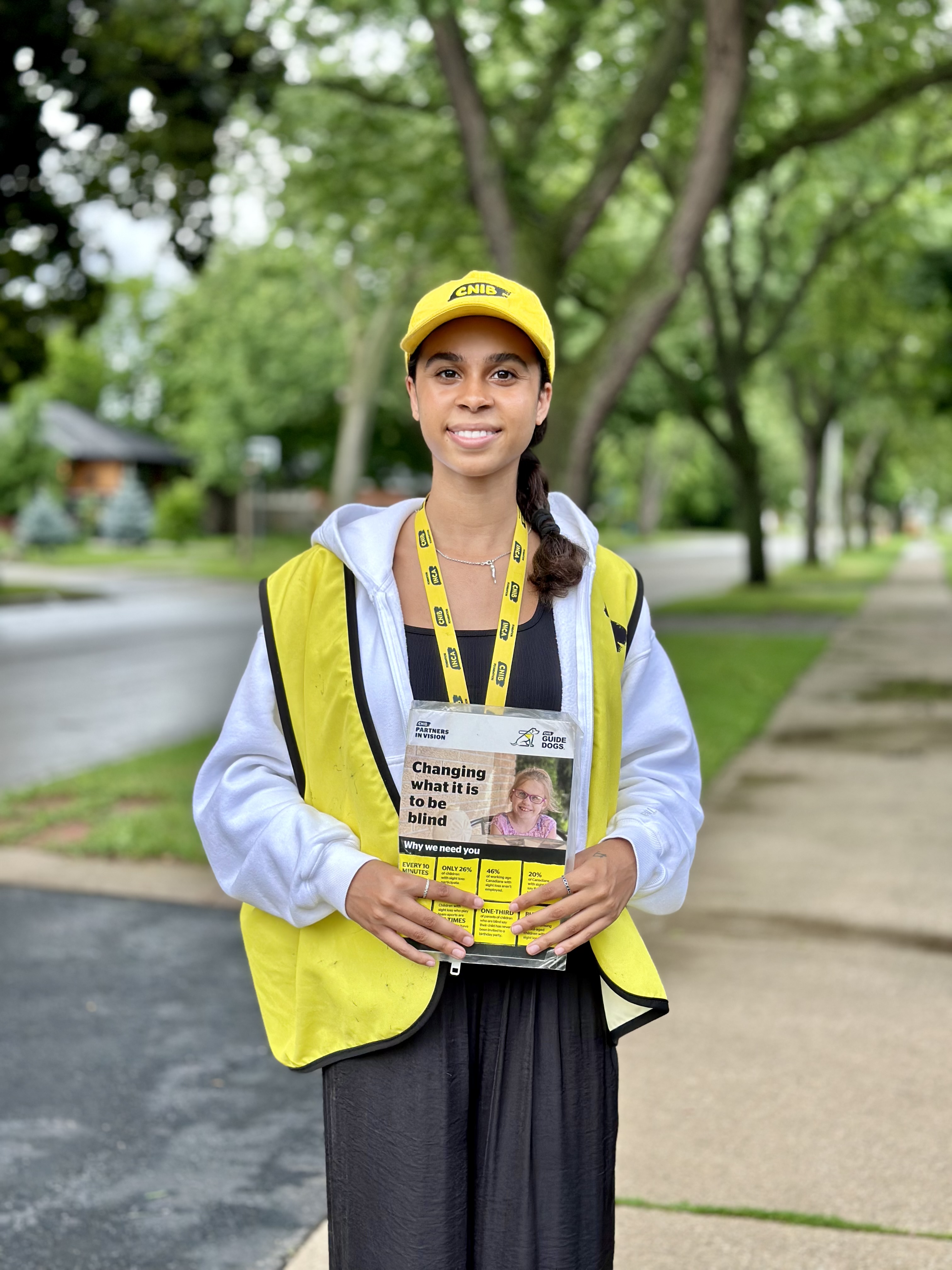 Un collecteur de fonds travaillant pour le compte d'INCA sourit à la caméra, debout sur un trottoir, sous des arbres verts. Elle porte une casquette jaune d'INCA, un cordon avec photo d'identité et un gilet, et tient une grande carte présentant des faits sur la cécité.