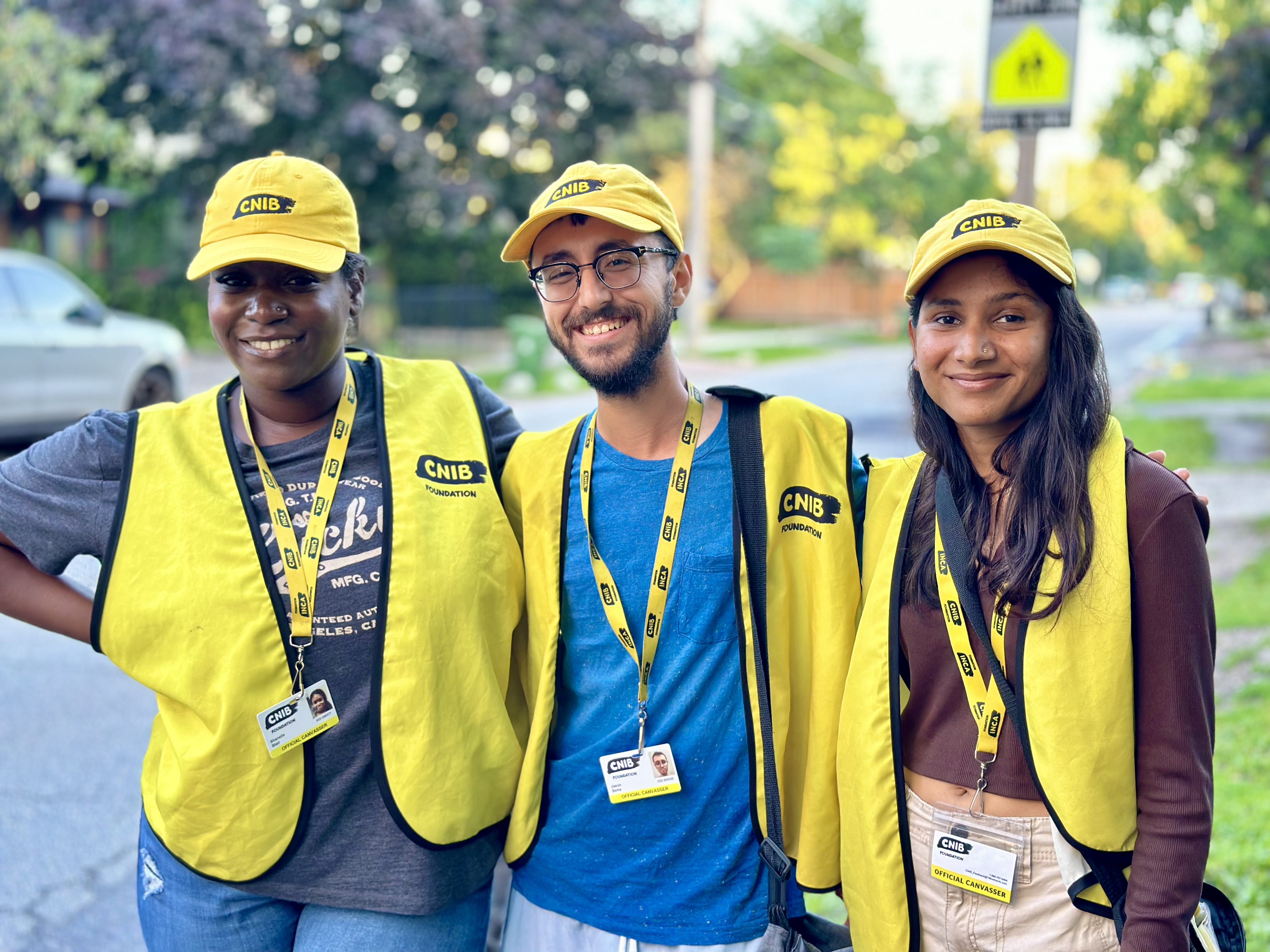 Trois collecteurs de fonds travaillant pour le compte d'INCA sourient à la caméra, debout sur le trottoir dans un quartier résidentiel. Chaque collecteur de fonds porte une casquette de baseball jaune d'INCA, un gilet et un cordon avec photo d'identité.