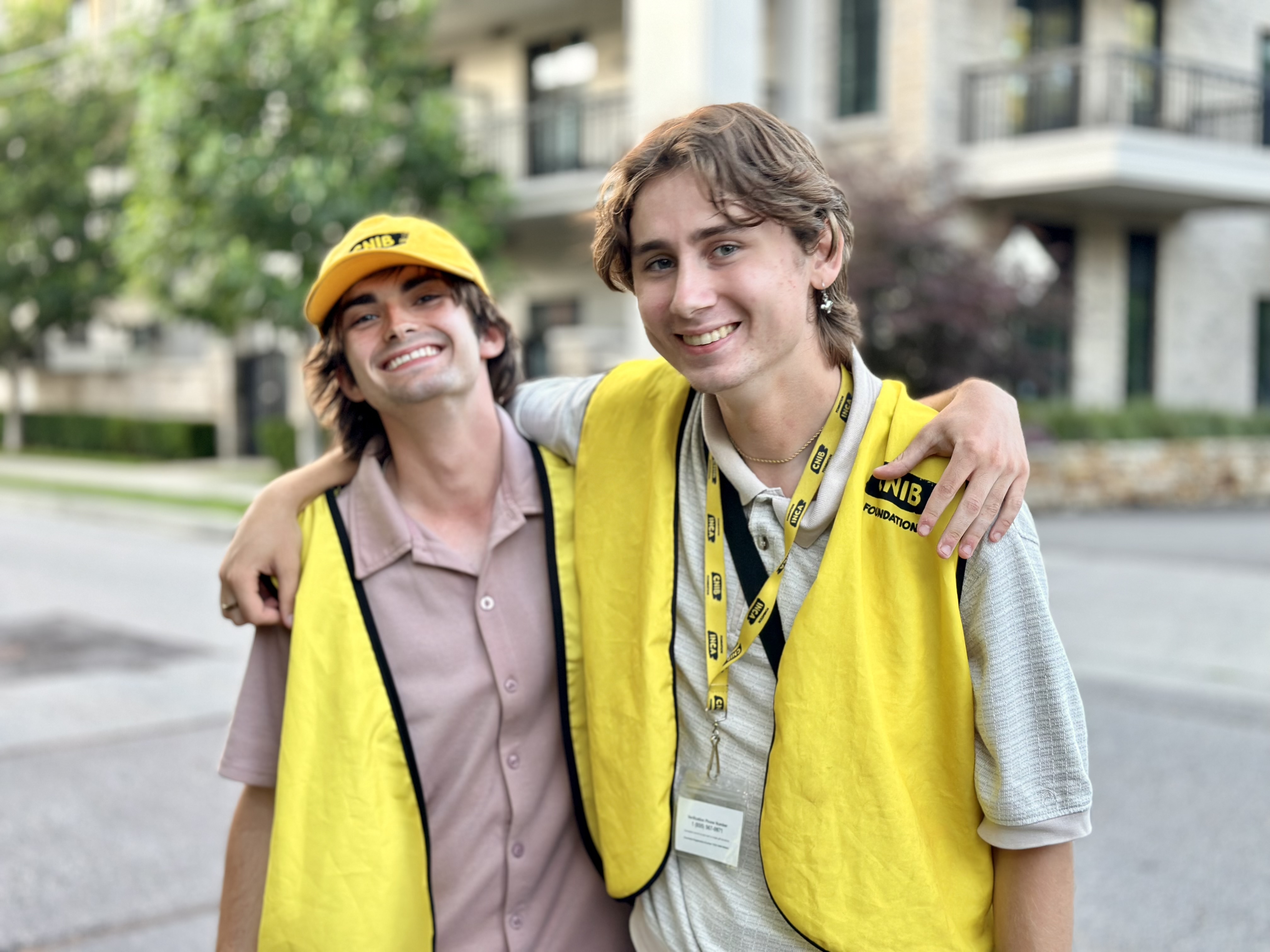 Deux collecteurs de fonds travaillant pour le compte d'INCA sourient en se tenant par le bras devant un immeuble d'habitation. Ils portent tous les deux un gilet jaune d'INCA. Celui de gauche porte aussi une casquette de baseball jaune d'INCA, tandis que celui de droite porte un cordon jaune.