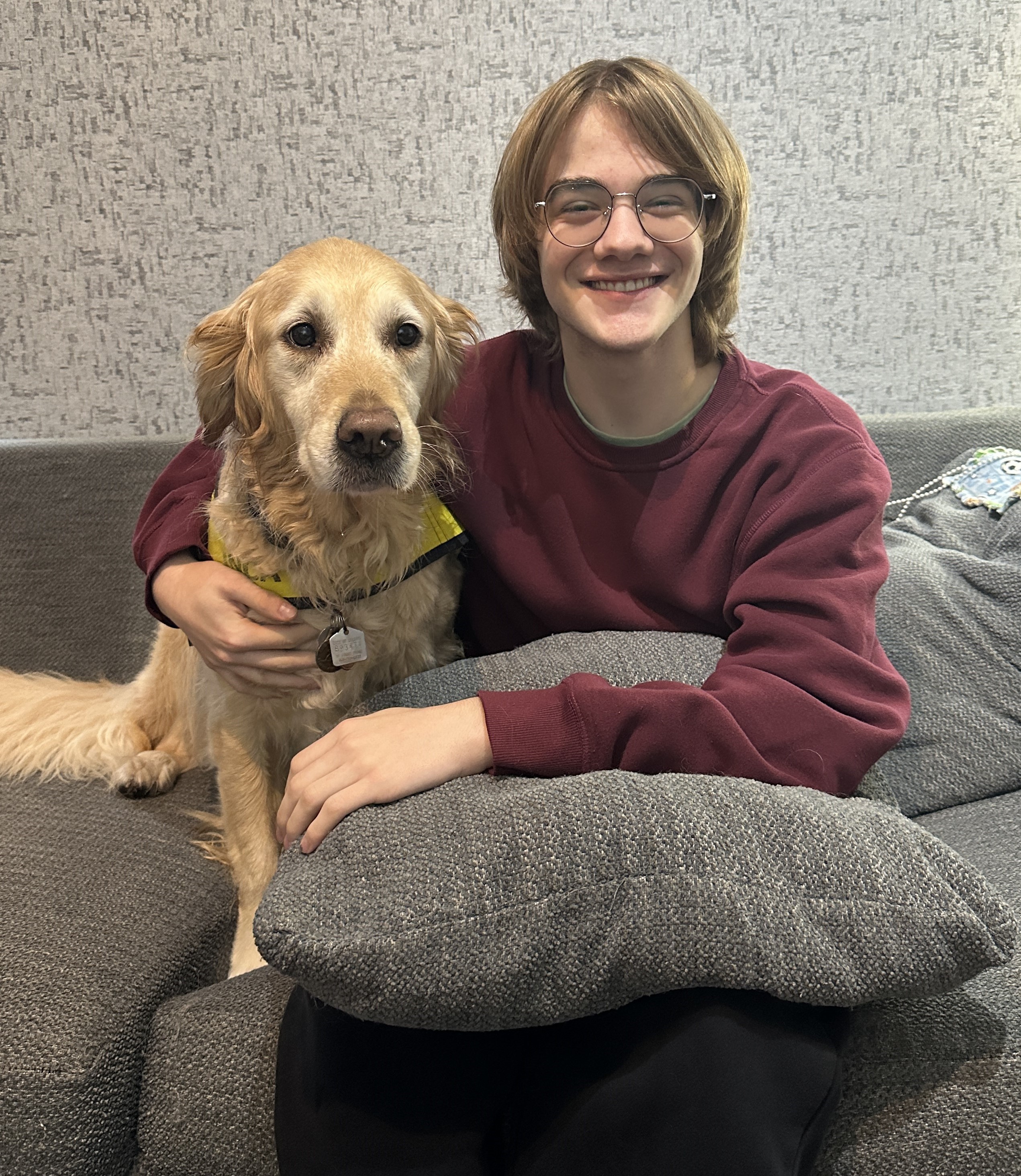 Gabriel sourit, assis sur un sofa avec Maggie, sa chienne compagne d’INCA au pelage blond et doux. Maggie porte une veste jaune d’INCA.