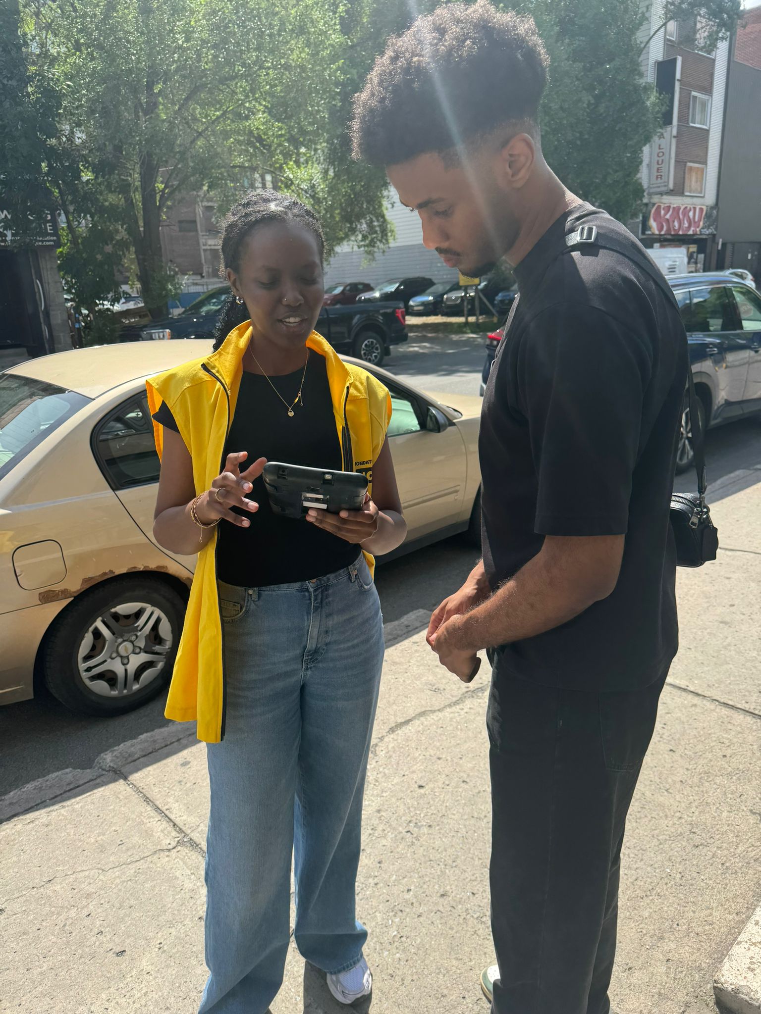 Une collectrice de fonds travaillant pour le compte d'INCA parle à une personne sur le trottoir tandis que toutes deux consultent l'écran de sa tablette sécurisée. La collectrice de fonds porte un gilet jaune d'INCA.