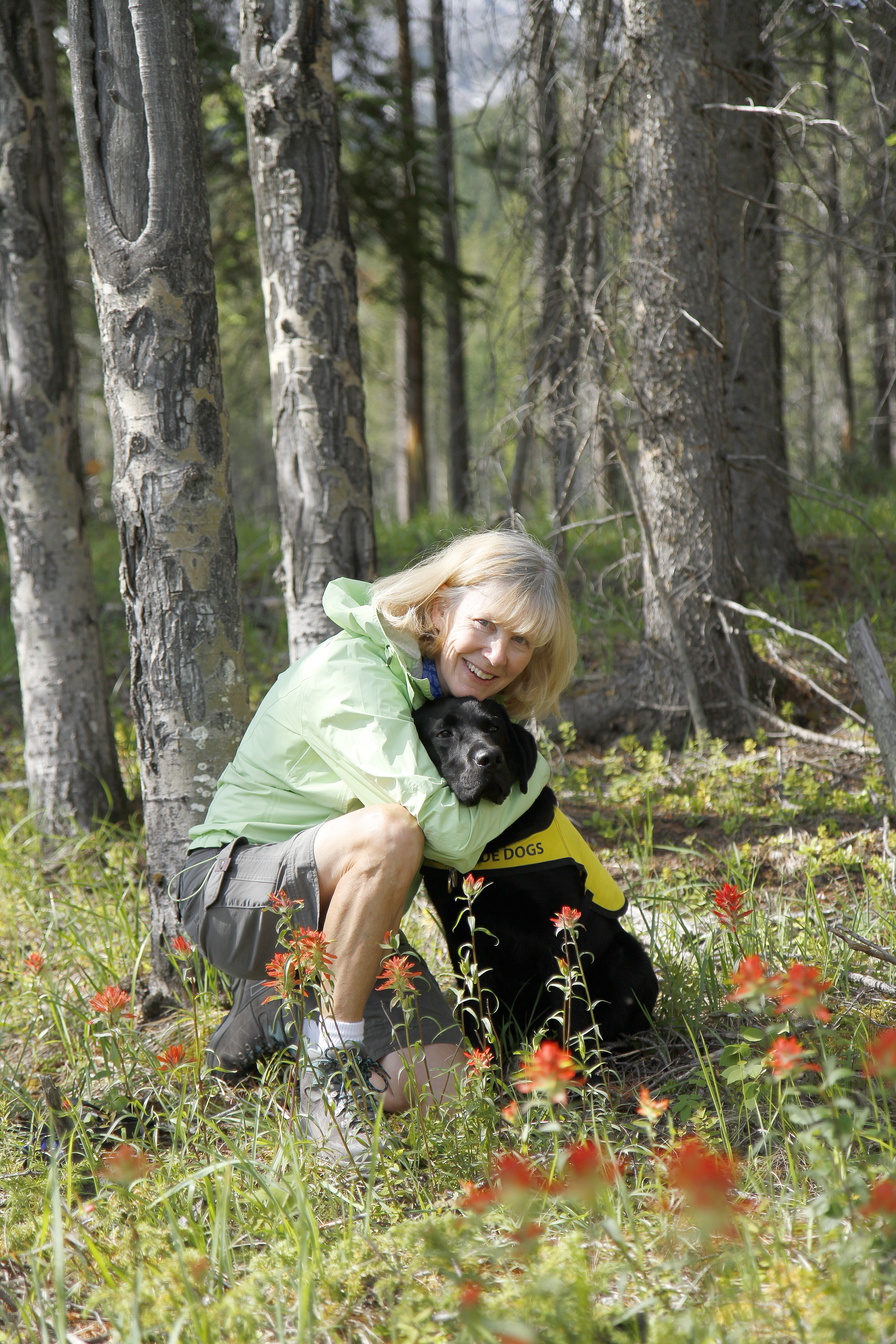 Cheryl kneeling next to Irwin, a black Labrador retriever wearing a yellow Future Guide Dog vest, in a forest in Canmore Alberta surrounded by trees and greenery.