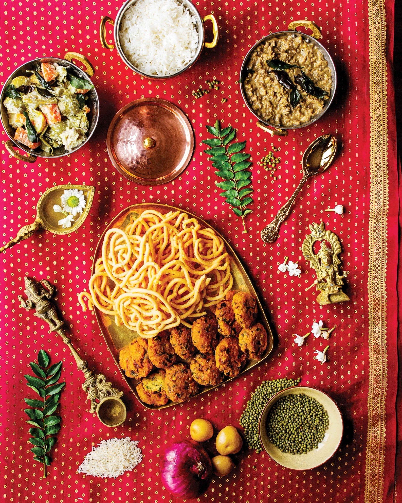 Plusieurs plats du livre, dont le murukku (casse-croûte croustillant), le vada (casse-croûte frit), l’avial (curry de noix de coco avec des légumes), le riz basmati et le dhal (lentilles), sont présentés dans des plats dorés sur un sari de soie magenta et or. Des ingrédients colorés, des fleurs de jasmin et une statue dorée se trouvent sur la table parmi les plats