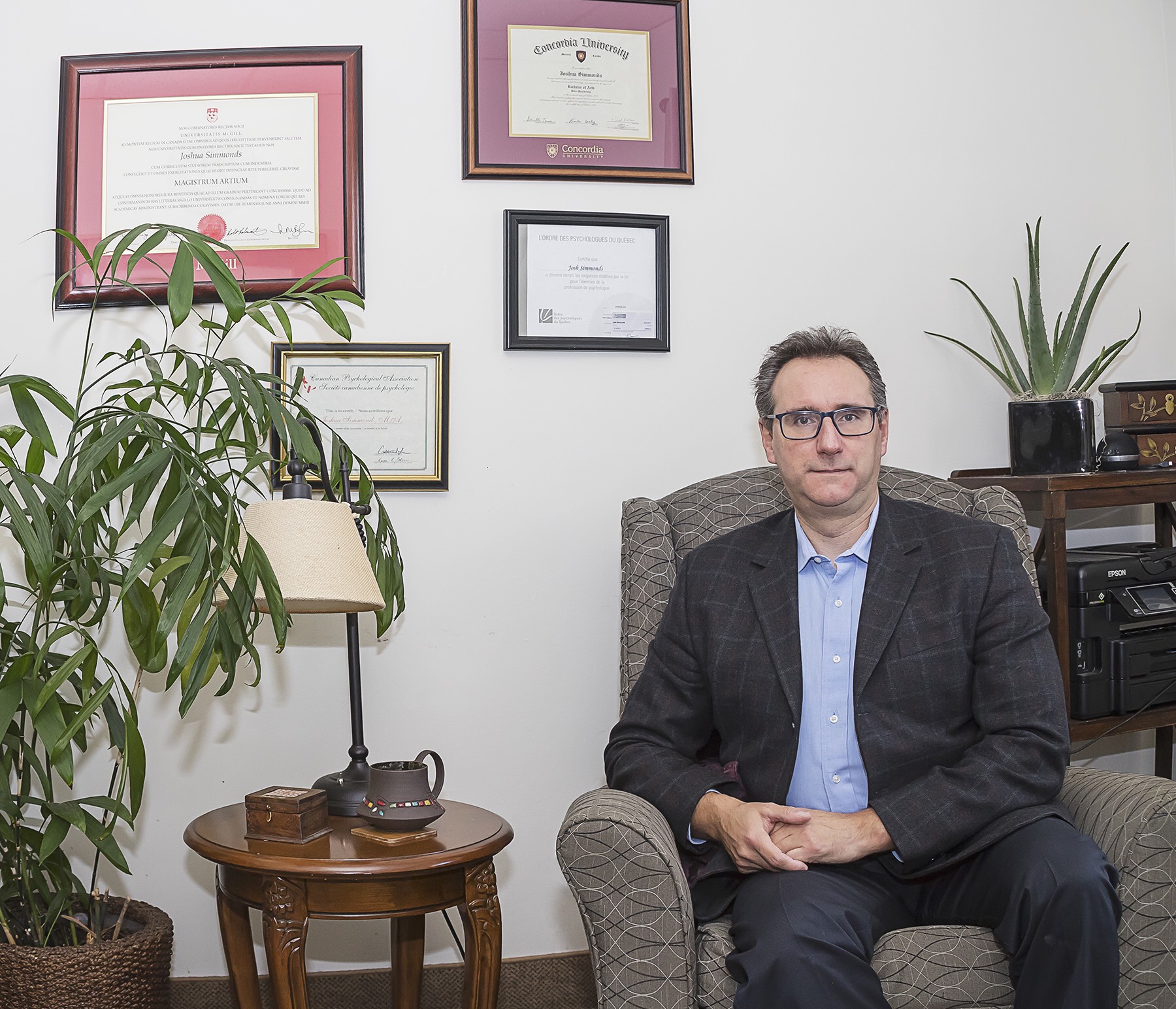 Joshua Simmonds, wearing a suit, sits in a chair in his office. There are certificates on the wall and plants and a coffee table beside him.