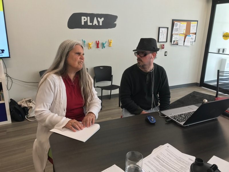 A man and woman sit at a table at the CNIB London Hub. She is reading a braille document. He has a laptop open in front of him.