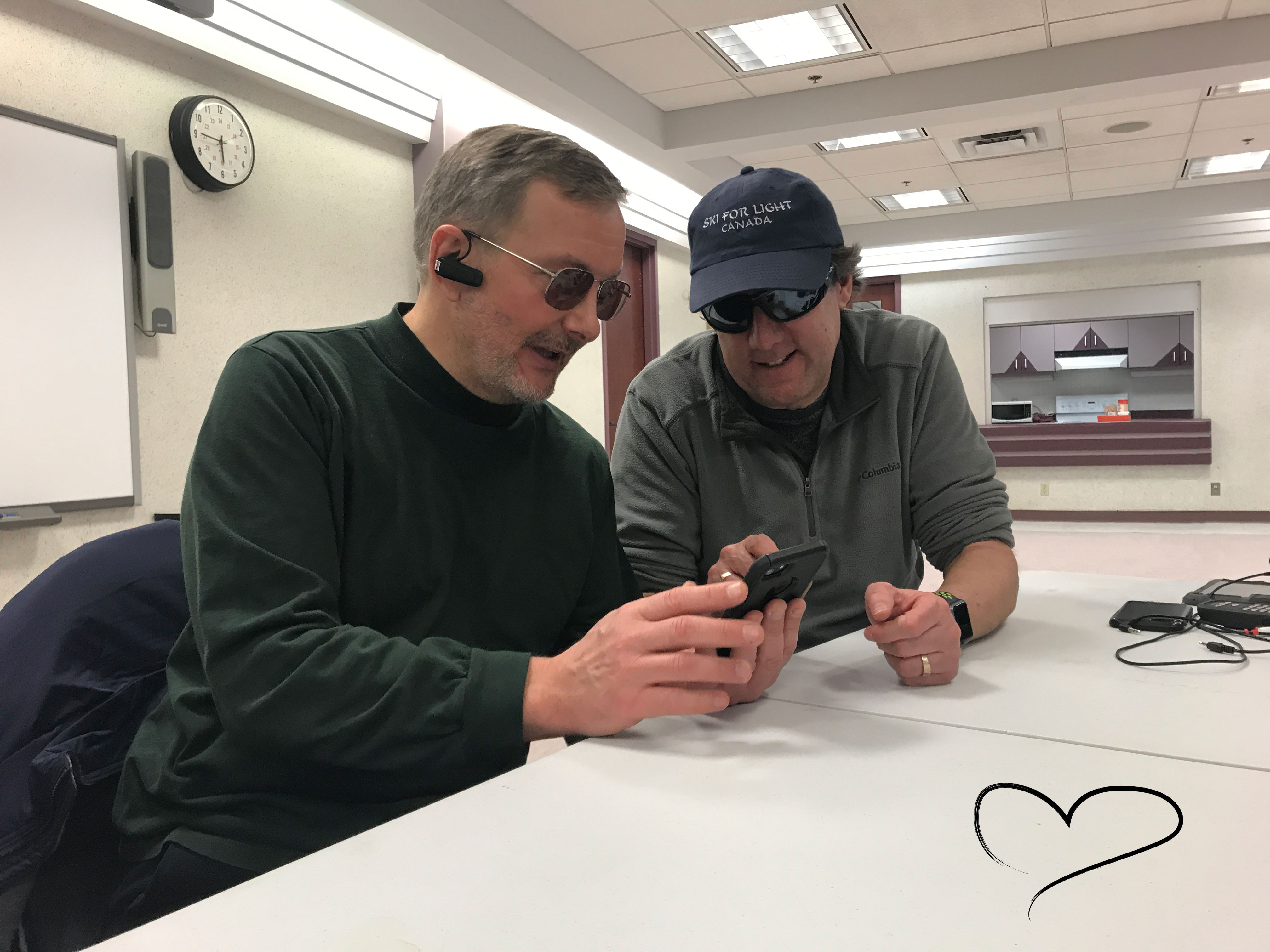 Lloyd Buckler, assis à une table, aidant quelqu'un à apprendre à utiliser son iPhone. Un dessin en forme de cœur se trouve sur la table, dans le coin inférieur droit de l'image.