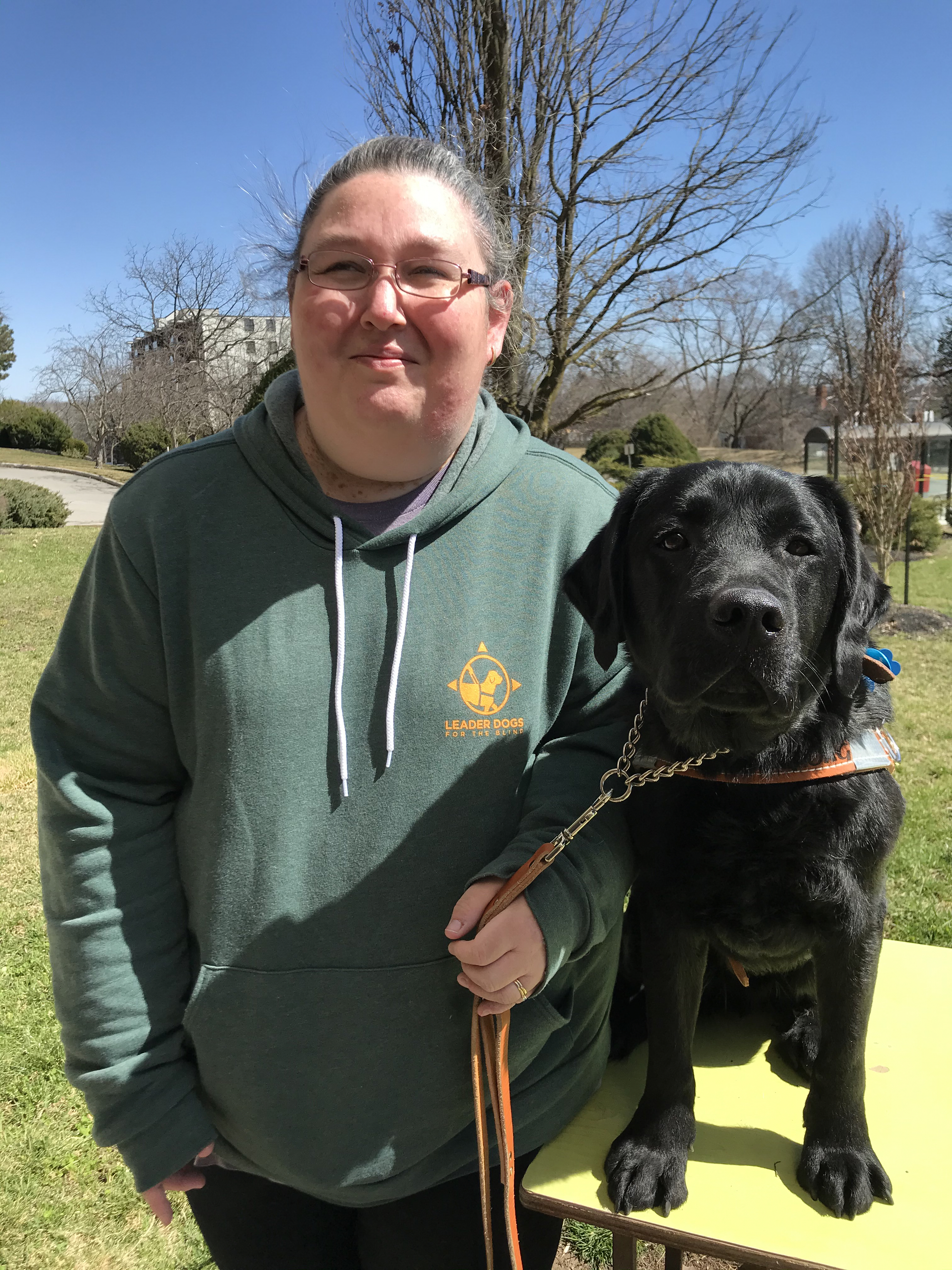 Lyn Morris and her guide dog, Leo, a black Labrador retriever, standing in a park and smiling for the camera