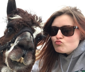Melyssa poses wearing sunglasses and pursing her lips beside her alpaca friend Lil Chiv.