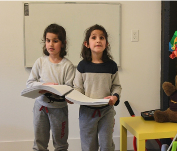Two young children are holding on to the same braille book and walking around a classroom environment.