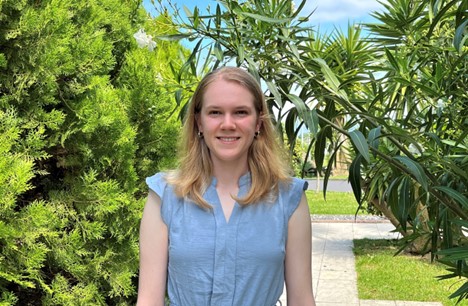 Laura, une jeune femme aux cheveux blonds, sourit devant un jardin verdoyant avec des palmiers et des arbustes. Elle porte une robe de soleil bleu clair.