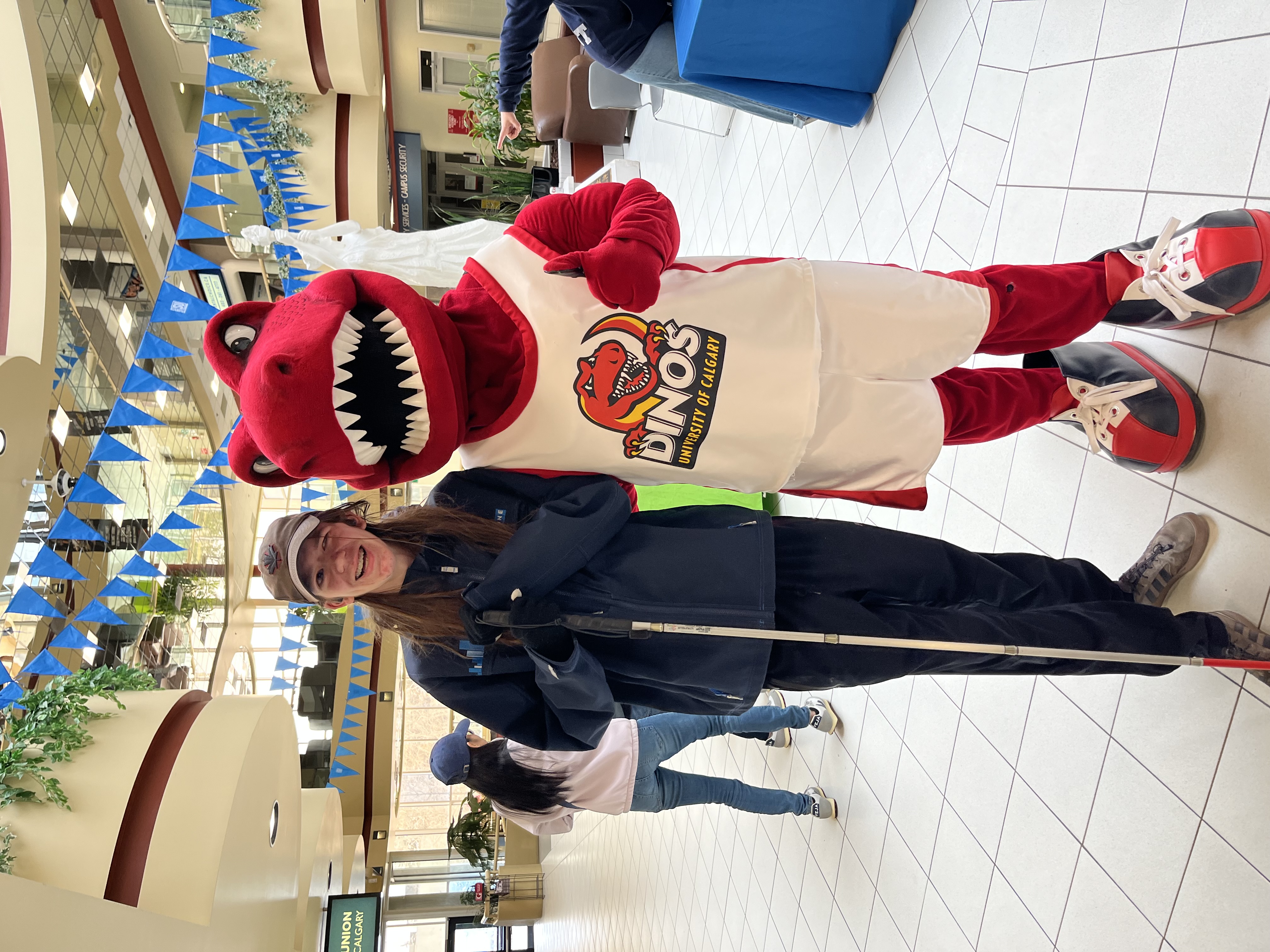 Zachary, un jeune homme aux longs cheveux bruns, sourit à côté de Rex, la mascotte dinosaure rouge et duveteuse de l'université de Calgary. Zach est vêtu d'un sweat-shirt bleu, d'un pantalon de survêtement, d'un chapeau et tient sa canne blanche.